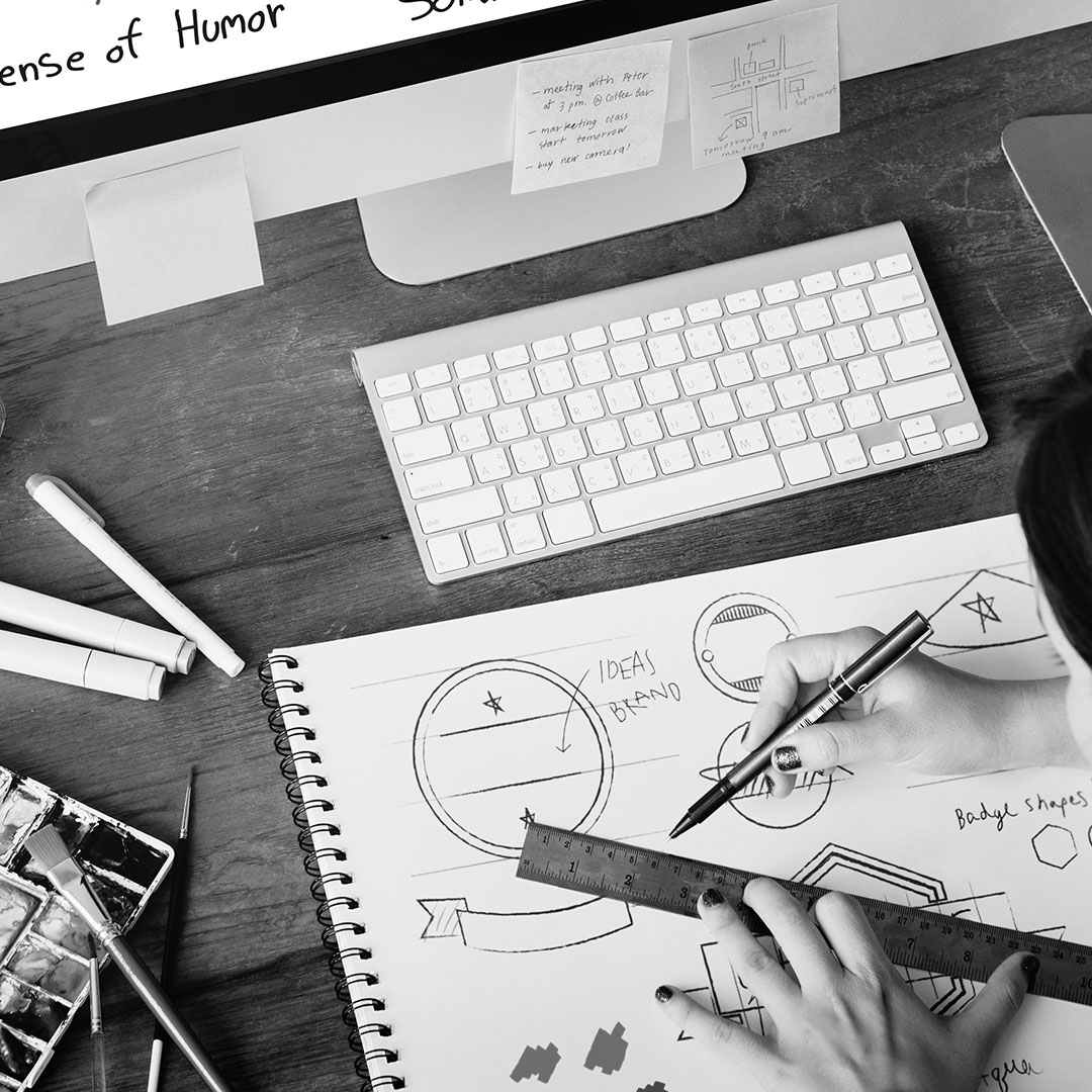 A person sketches logos in a book, surrounded by various design elements on their workspace.